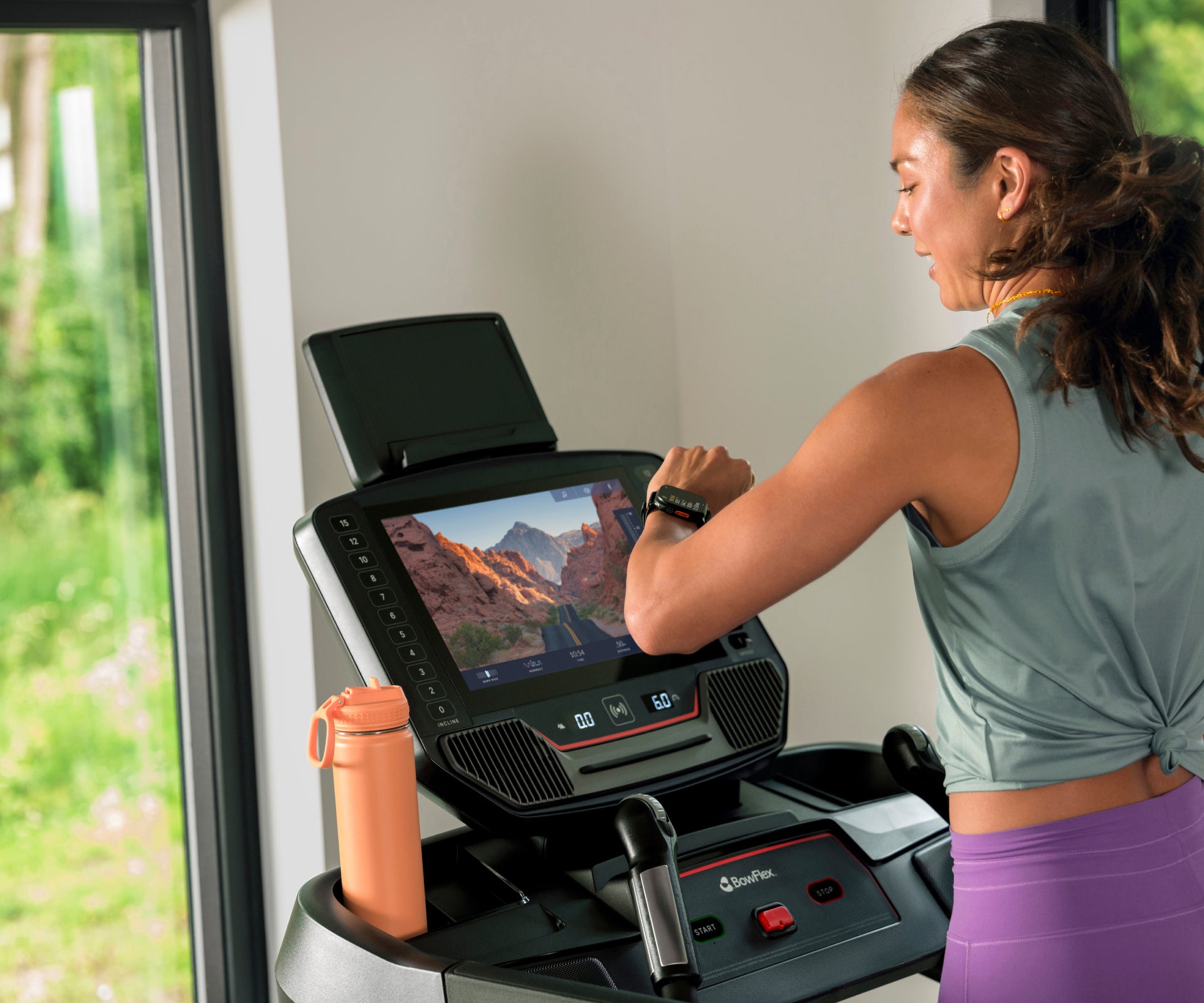 Woman using the BowFlex T16 Treadmill with a JRNY app screen displayed on its touchscreen console, with a water bottle in the treadmill's water bottle holder.