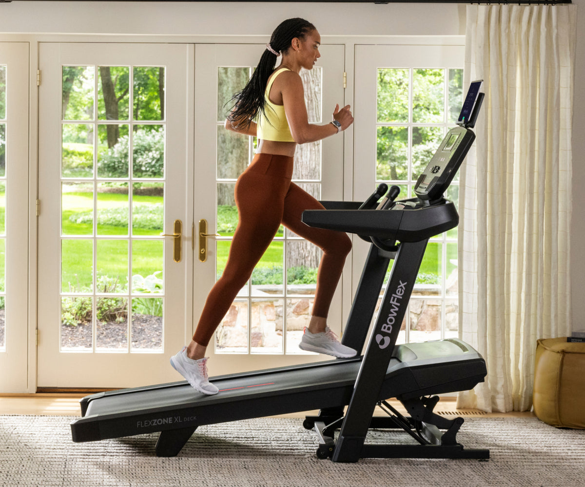 A woman in athletic wear running on a BowFlex T9 Treadmill in a bright room with large windows, cream curtains, and a light rug.