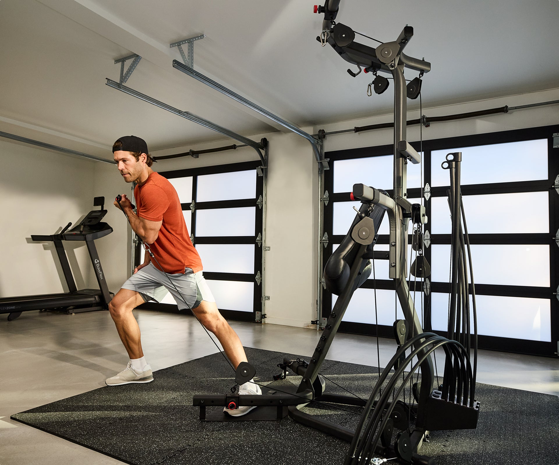 A man in an orange t-shirt and gray shorts working out using the cable pulley system on the BowFlex Xtreme 2 SE Home Gym in a modern home gym with garage doors and a BowFlex treadmill in the background.
