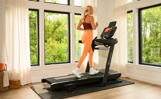 A woman in athletic wear runs on a BowFlex T16 Treadmill in a bright room with large windows and green outdoor views.