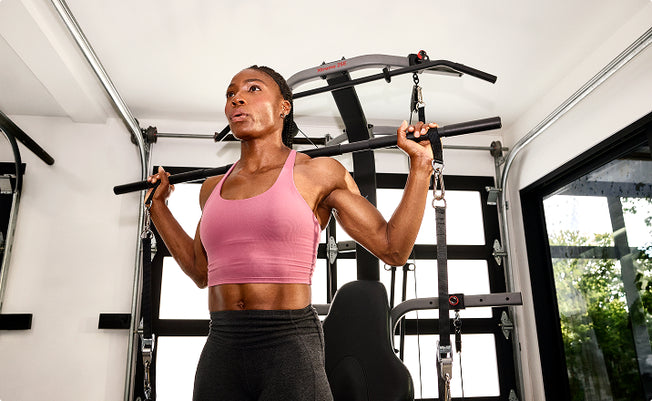 A woman in a pink sports bra and dark gray leggings working out using the 6-grip steel straight bar on the BowFlex Xtreme 2 SE Home Gym inside a room with white walls and large windows .