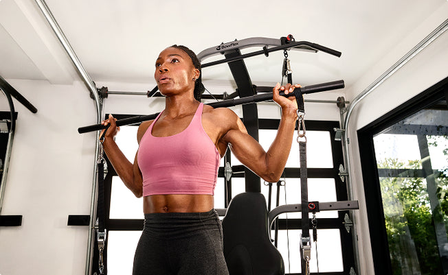 A woman in a pink sports bra and dark gray leggings working out using the 6-grip steel straight bar on the BowFlex Xtreme 2 SE Home Gym inside a room with white walls and large windows.