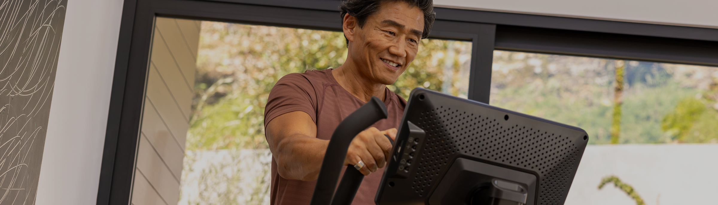Man smiling while exercising on the BowFlex Max Total 16 in a home setting with a blurred outdoor background visible through the windows.