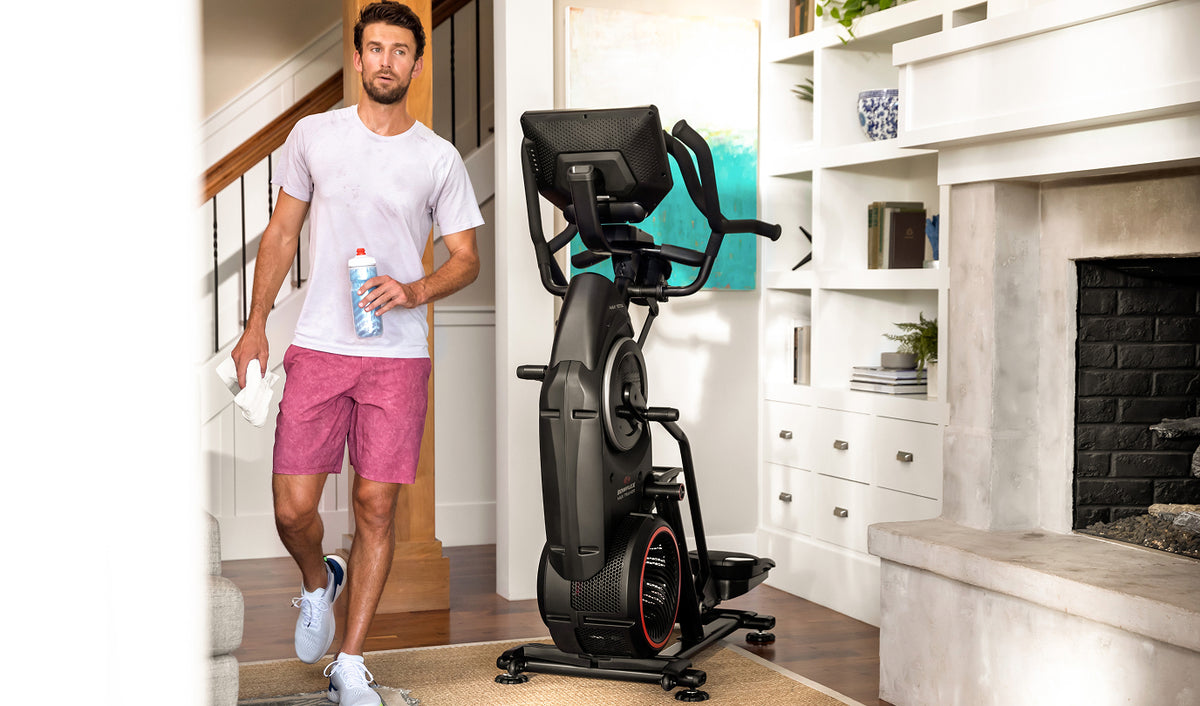 A man in a white t-shirt and pink shorts walks past the BowFlex Max Total 16 in a modern living room with a fireplace and white shelves.