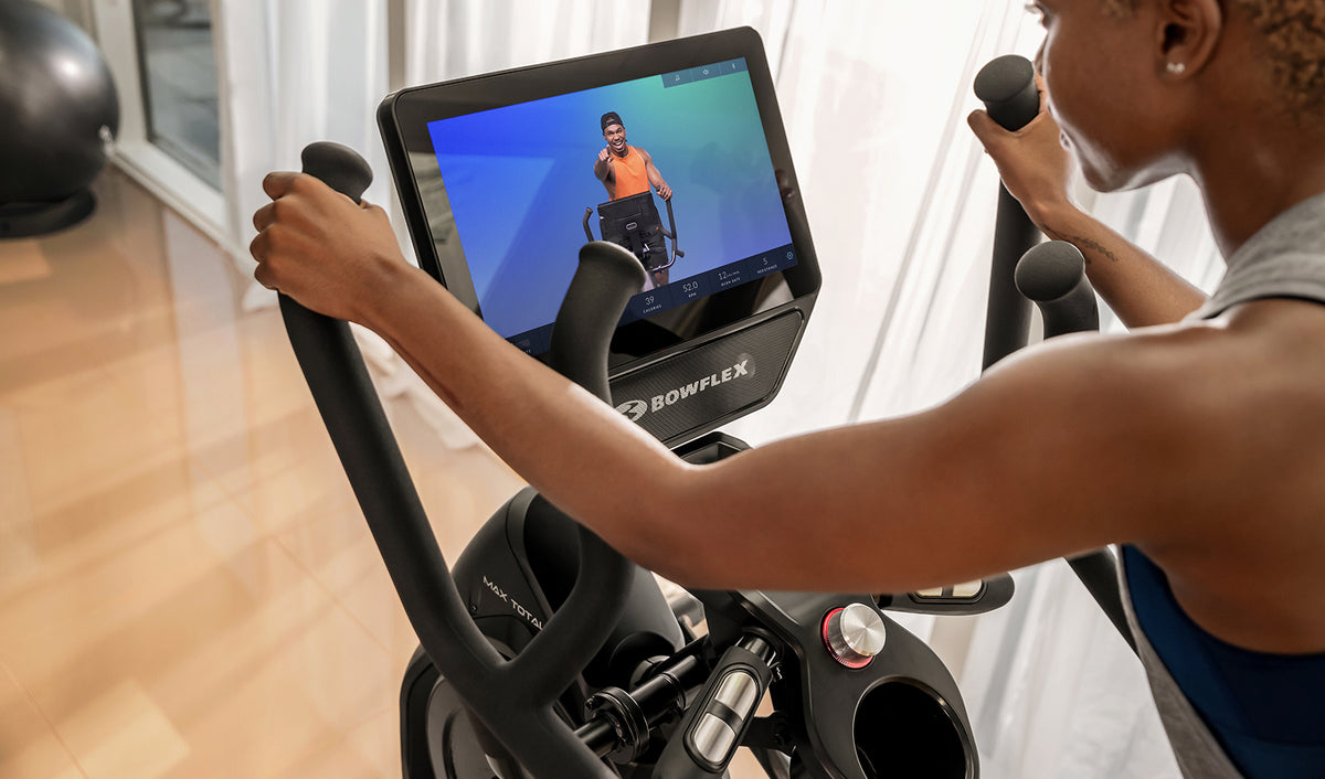A close-up of a person using the BowFlex Max Total 16 showing a JRNY app screen on its touchscreen. The person is seen from behind, holding the handlebars in a bright, modern indoor setting.