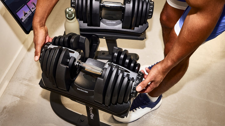 A close-up of a person adjusting a Results Series 1090 SelectTech Dumbbell on a dumbbell stand in a home gym setting. 