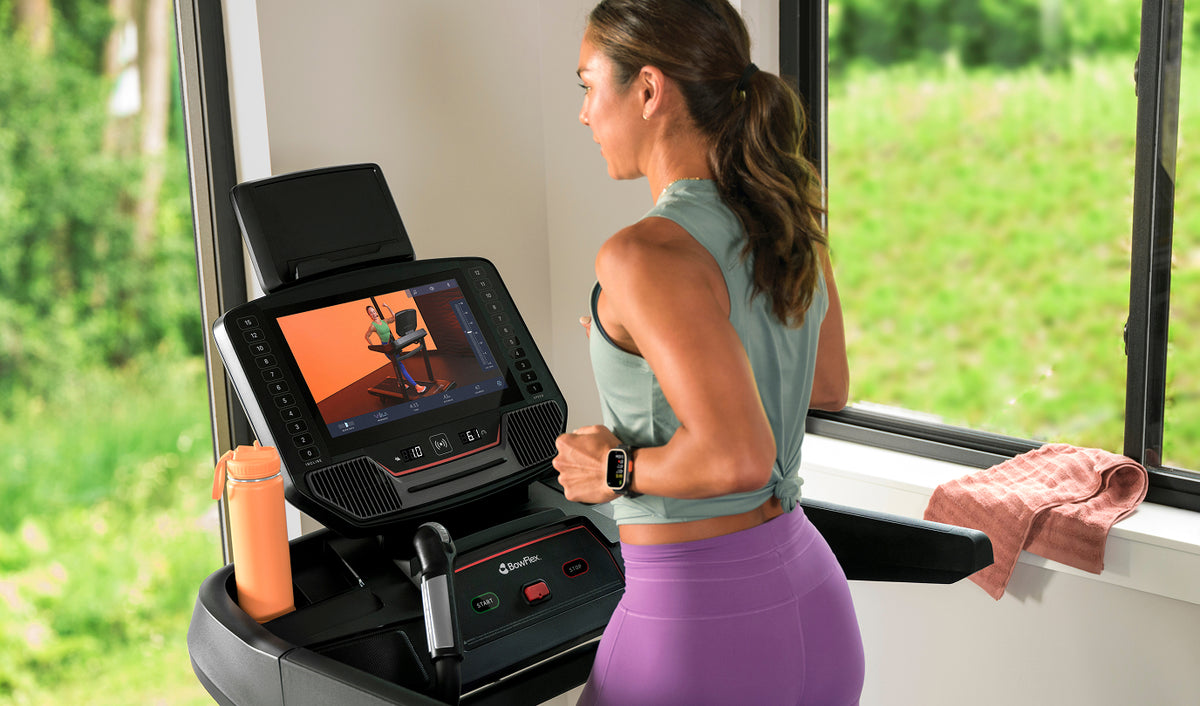 A woman in athletic wear runs on a BowFlex T16 Treadmill in a bright room with large windows and green outdoor views with a JRNY app screen displayed on the touchscreen console.