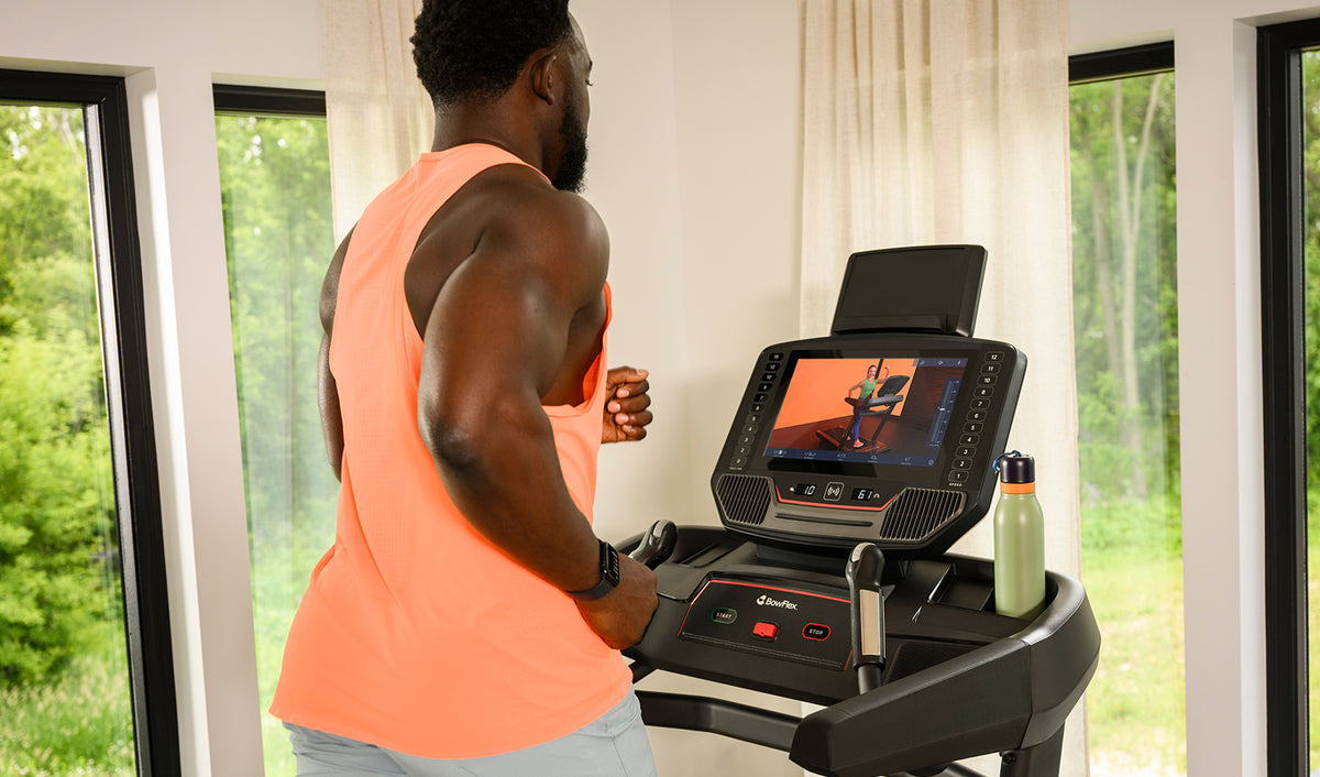 A man in athletic wear runs on a BowFlex T16 Treadmill in a bright room with large windows and green outdoor views.