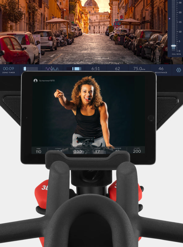 A tablet on the BowFlex VeloCore Bike 22 media rack displaying a JRNY app screen. The product console is just behind the tablet.
