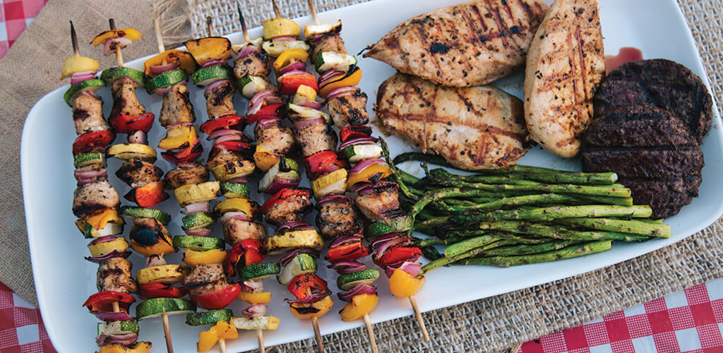 A plate of chicken and veggie kabobs next to grilled chicken, burgers, and asparagus.