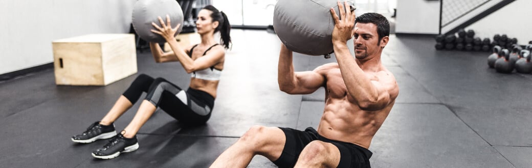 Two fit people in a gym performing one of the best abs workout with a medicine ball. Ab Workout Guide – Ab-solutely the Best Ab Exercises.