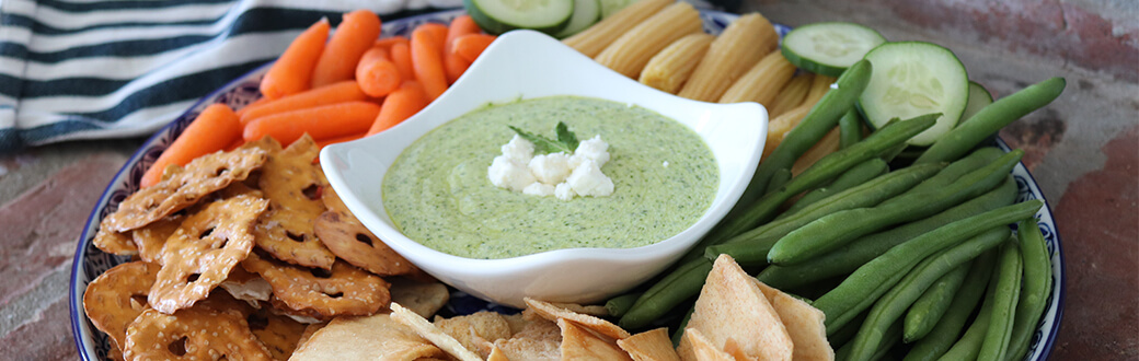 A plate of crackers and chopped veggies surrounding a bowl of Greek dip.