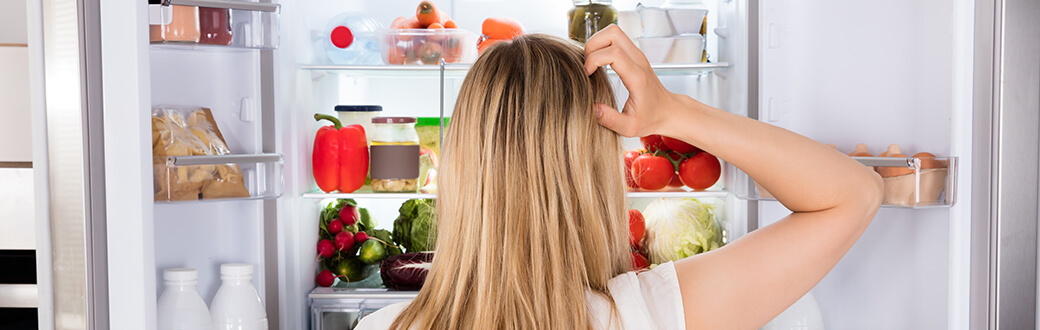 A woman scratching her head in front of an open refrigerator.