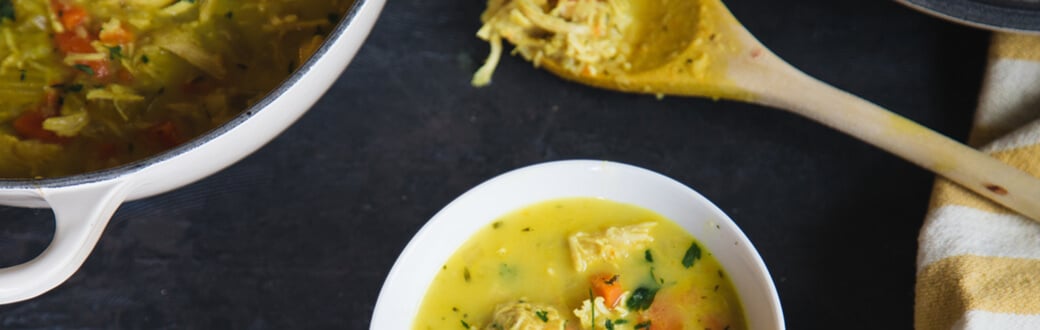 A pot and bowl of chicken soup next to a wooden spoon on a table.