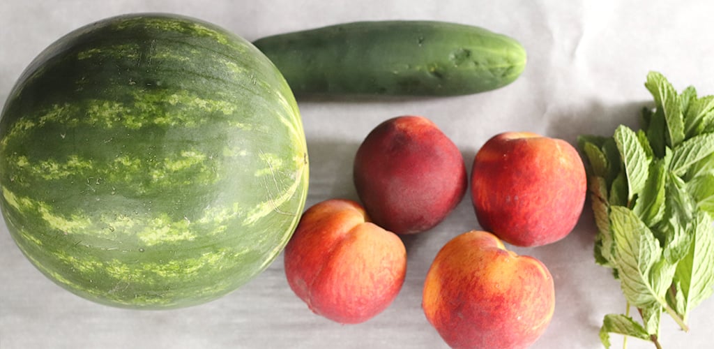 Watermelon, cucumber, peaches, and mint on a cutting board.